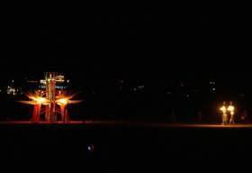 Two lighted figures walk to the wooden flower
