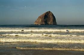 Two men take out Sea Kayaks to go surfing in them, before a rock with arch.